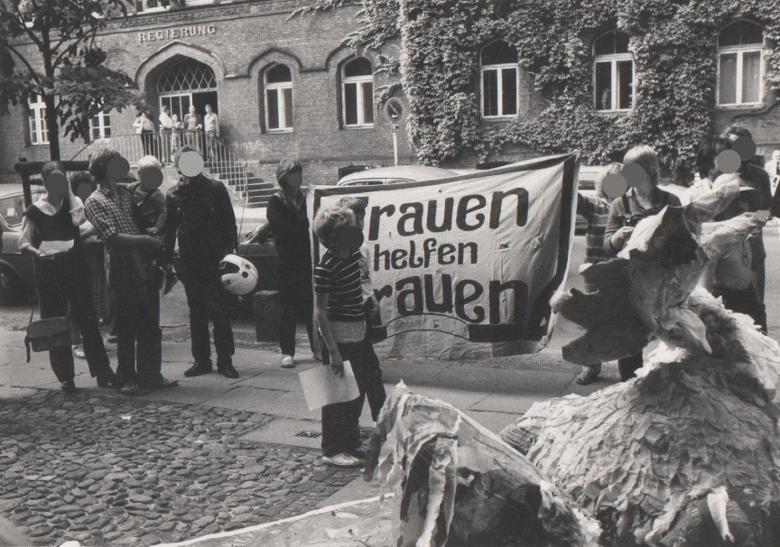 Auf dem Bild ist eine Gruppe von Menschen zu sehen, die vor dem Lüneburger Rathaus stehen. Zwei Menschen halten ein Transparent. Auf dem steht: Frauen helfen Frauen. Das Foto ist vermutlich 1984 entstanden.